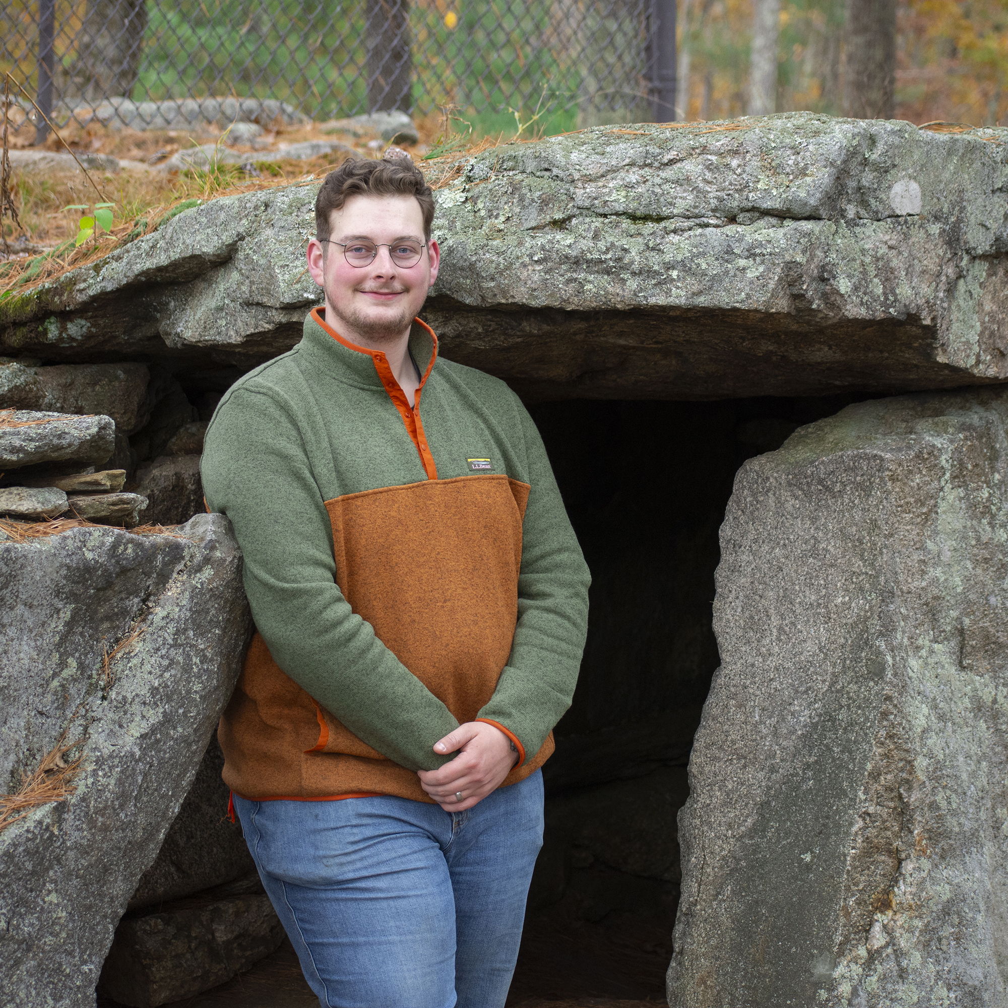 A casually-dressed man in blue jeans and a flannel sweater is relaxed as he leans against the large stone chamber next to him, which rivals his height. A gentle smile crosses his face.